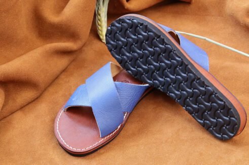 A pair of handmade Moroccan sandals featuring vibrant blue leather cross straps, brown stitched leather soles, and a natural beige leather lining, placed on a camel suede background with wheat stems.