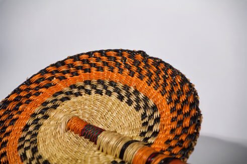 A handcrafted Moroccan fan with vibrant orange, black, and beige woven design, complemented by a leather handle, placed on a rocky surface with a scenic background