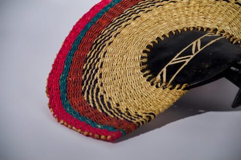 A handcrafted Moroccan fan featuring a colorful woven design with pink, green, and red tones, complemented by a wooden handle, placed on a rocky surface with a clear sky in the background.