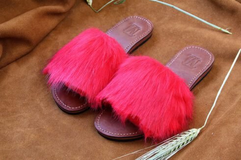 A pair of handmade Moroccan slide sandals featuring vibrant red hair-on leather uppers and brown leather soles, laid on a camel suede background with natural wheat details.