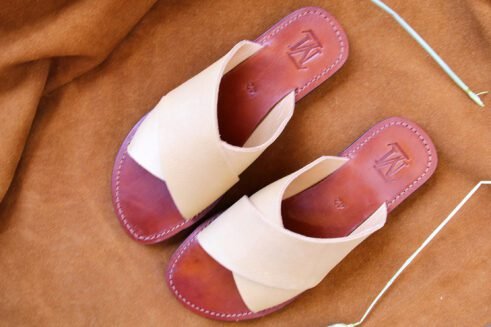 A pair of handmade Moroccan leather slide sandals with beige cross-over straps and brown leather soles, styled on a camel suede background with wheat accents.