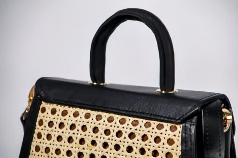 Chic handbag with a black leather top and woven cane body, featuring a gold-tone clasp, a sturdy handle, and an adjustable shoulder strap, styled against a soft neutral backdrop.