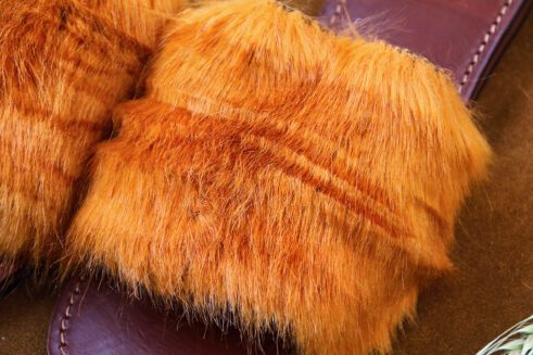 A pair of handmade Moroccan slide sandals featuring a wide burnt orange hair-on leather upper and brown stitched leather footbeds, set against a camel suede background with wheat accents