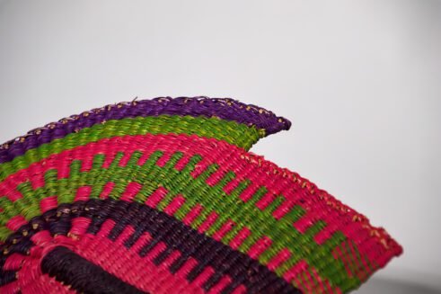 A handcrafted Moroccan fan with vibrant pink, green, and purple colors, complemented by a leather handle, placed on a rocky surface with a scenic background.