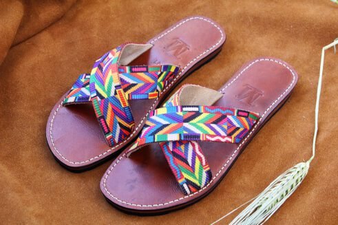 A pair of handmade Moroccan sandals featuring multicolor ethnic-patterned woven fabric in a cross-strap design over brown leather soles, displayed on a camel suede background with wheat accents