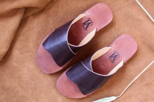 A pair of handmade Moroccan sandals featuring wide dark brown leather cross straps and stacked leather soles with a black rubber base, set on a camel suede background with wheat decor