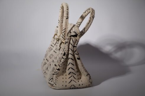 A handcrafted Moroccan tote bag with a tribal black geometric design on beige fabric, placed on a neutral background surrounded by small stones.