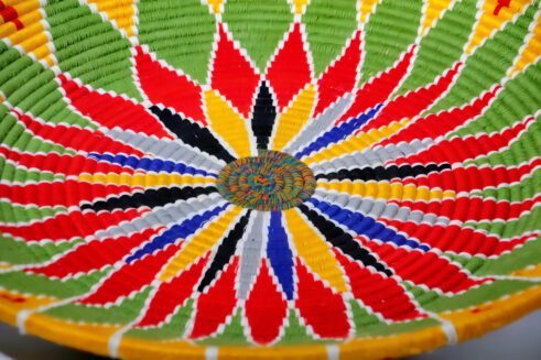A beautifully handcrafted Moroccan coil basket featuring a bold flower design in red, yellow, green, blue, black, and white colors, set against a neutral background with small stones nearby.