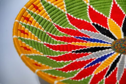 A beautifully handcrafted Moroccan coil basket featuring a bold flower design in red, yellow, green, blue, black, and white colors, set against a neutral background with small stones nearby.