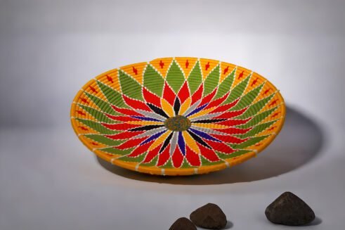 A beautifully handcrafted Moroccan coil basket featuring a bold flower design in red, yellow, green, blue, black, and white colors, set against a neutral background with small stones nearby.