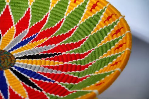 A beautifully handcrafted Moroccan coil basket featuring a bold flower design in red, yellow, green, blue, black, and white colors, set against a neutral background with small stones nearby.