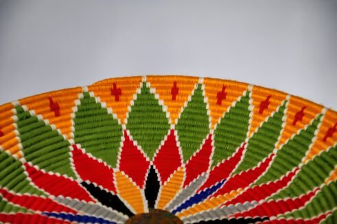 A beautifully handcrafted Moroccan coil basket featuring a bold flower design in red, yellow, green, blue, black, and white colors, set against a neutral background with small stones nearby.