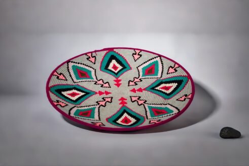 A beautifully handcrafted Moroccan coil basket with a geometric design featuring pink, teal, black, and white colors, placed on a neutral background with a small stone beside it.