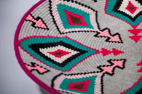 A beautifully handcrafted Moroccan coil basket with a geometric design featuring pink, teal, black, and white colors, placed on a neutral background with a small stone beside it.