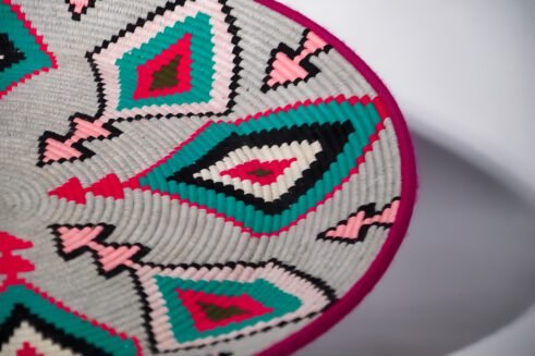 A beautifully handcrafted Moroccan coil basket with a geometric design featuring pink, teal, black, and white colors, placed on a neutral background with a small stone beside it.