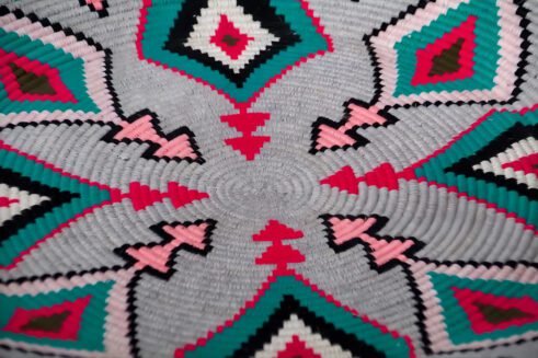 A beautifully handcrafted Moroccan coil basket with a geometric design featuring pink, teal, black, and white colors, placed on a neutral background with a small stone beside it.