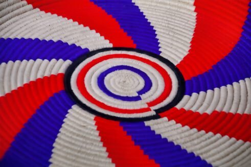 A handcrafted Moroccan coil basket featuring a red, blue, and beige spiral design, placed on a neutral background with small stones surrounding it.