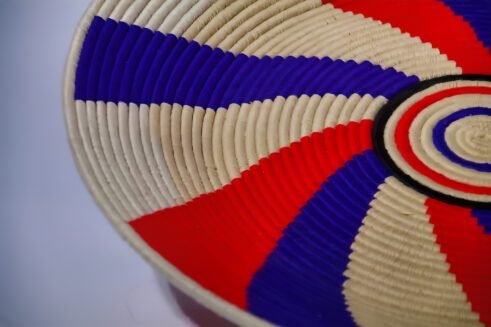A handcrafted Moroccan coil basket featuring a red, blue, and beige spiral design, placed on a neutral background with small stones surrounding it.