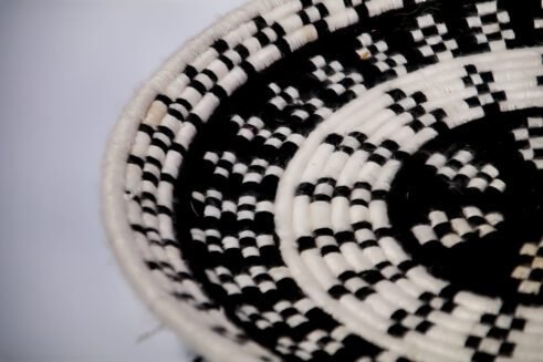 A handcrafted Moroccan coil basket with a geometric black and white design, placed on a neutral background with small stones surrounding it.
