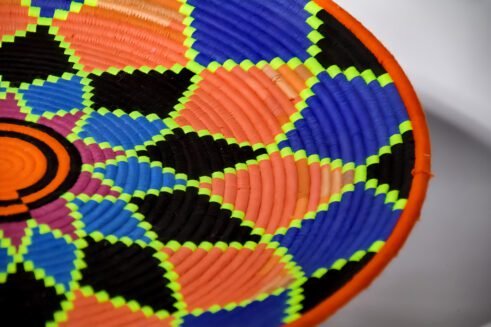 A handcrafted Moroccan coil basket featuring a vibrant geometric flower design in neon orange, pink, blue, and green, placed on a neutral background surrounded by small stones.