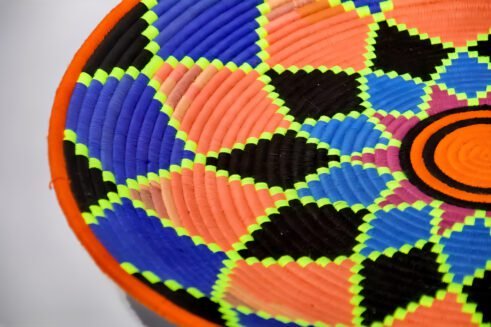 A handcrafted Moroccan coil basket featuring a vibrant geometric flower design in neon orange, pink, blue, and green, placed on a neutral background surrounded by small stones.
