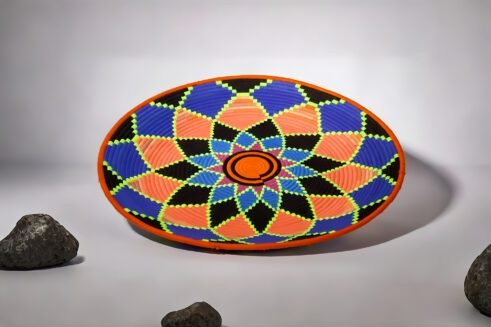 A handcrafted Moroccan coil basket featuring a vibrant geometric flower design in neon orange, pink, blue, and green, placed on a neutral background surrounded by small stones.