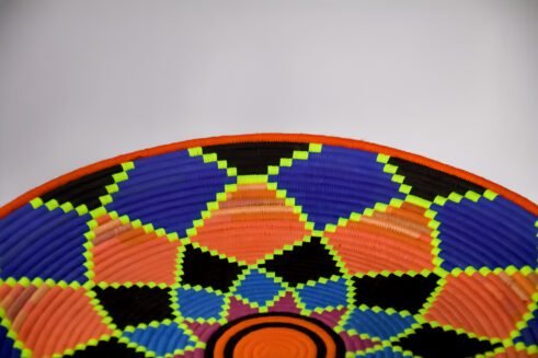 A handcrafted Moroccan coil basket featuring a vibrant geometric flower design in neon orange, pink, blue, and green, placed on a neutral background surrounded by small stones.