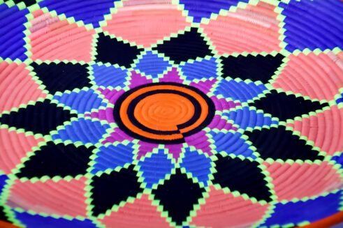 A handcrafted Moroccan coil basket featuring a vibrant geometric flower design in neon orange, pink, blue, and green, placed on a neutral background surrounded by small stones.
