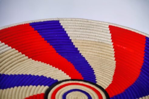 A handcrafted Moroccan coil basket featuring a red, blue, and beige spiral design, placed on a neutral background with small stones surrounding it.