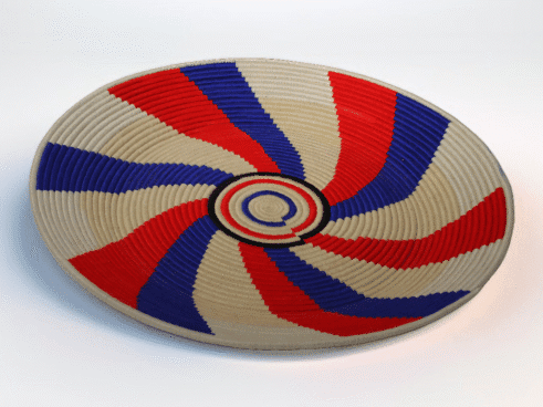 A handcrafted Moroccan coil basket featuring a red, blue, and beige spiral design, placed on a neutral background with small stones surrounding it.