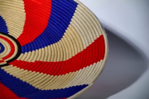 A handcrafted Moroccan coil basket featuring a red, blue, and beige spiral design, placed on a neutral background with small stones surrounding it.