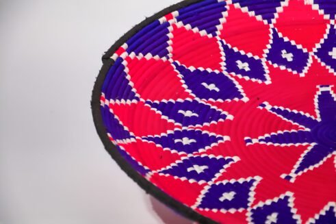 A handcrafted Moroccan coil basket with a vibrant pink and blue star design, surrounded by small stones on a neutral background.