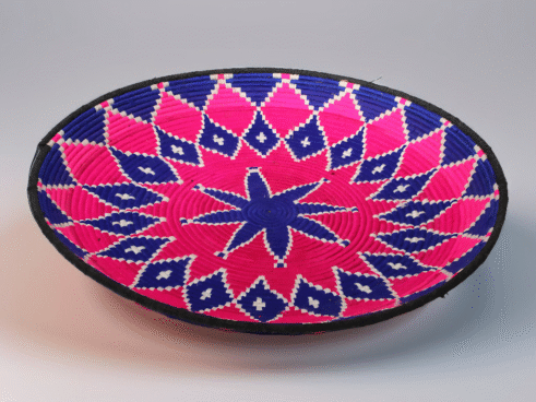 A handcrafted Moroccan coil basket with a vibrant pink and blue star design, surrounded by small stones on a neutral background.