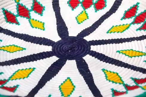 A handcrafted Moroccan coil basket with a colorful geometric design in green, yellow, red, and black, placed on a neutral background with small stones surrounding it.