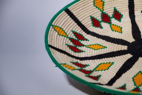 A handcrafted Moroccan coil basket with a colorful geometric design in green, yellow, red, and black, placed on a neutral background with small stones surrounding it.