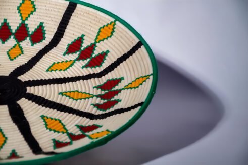 A handcrafted Moroccan coil basket with a colorful geometric design in green, yellow, red, and black, placed on a neutral background with small stones surrounding it.