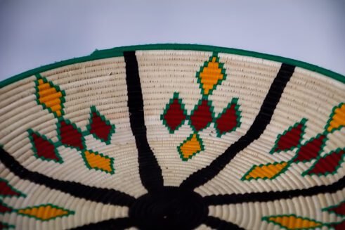A handcrafted Moroccan coil basket with a colorful geometric design in green, yellow, red, and black, placed on a neutral background with small stones surrounding it.