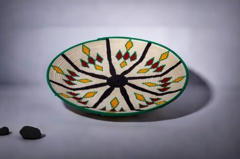 A handcrafted Moroccan coil basket with a colorful geometric design in green, yellow, red, and black, placed on a neutral background with small stones surrounding it.